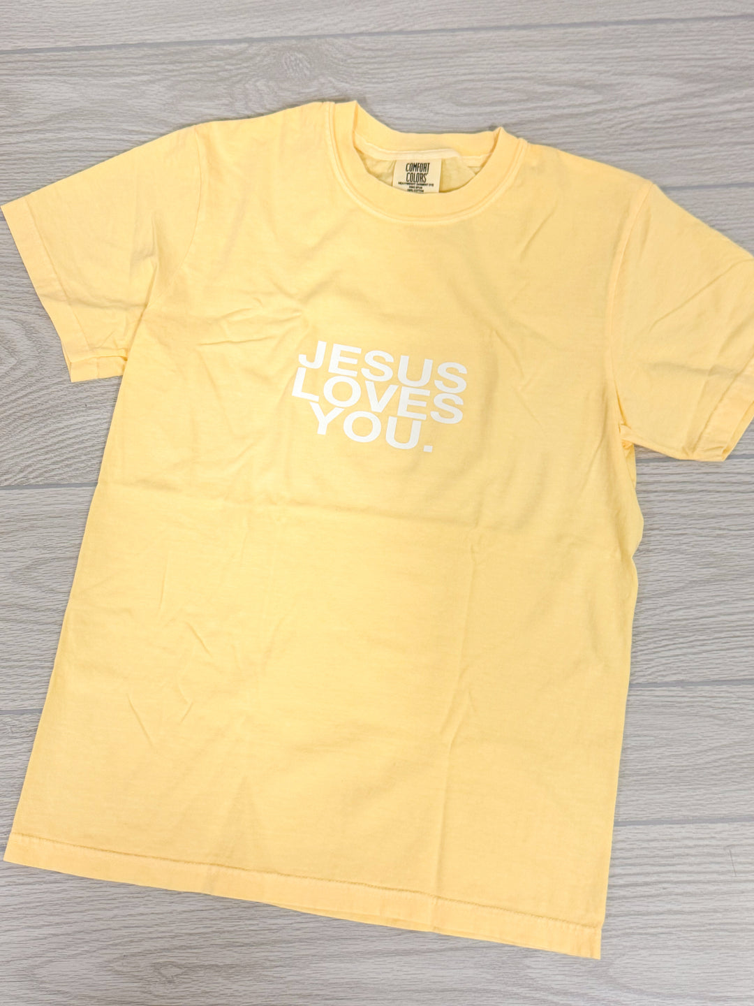 Yellow t-shirt with 'Jesus Loves You' text on a gray wooden surface