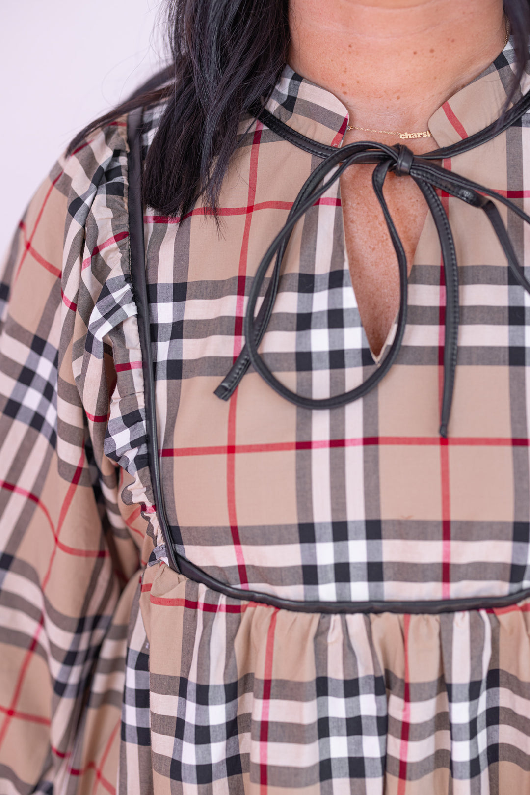 Women's taupe, black and red plaid babydoll dress with faux leather trim by Jodifl showing v neckline with adjustable self tie