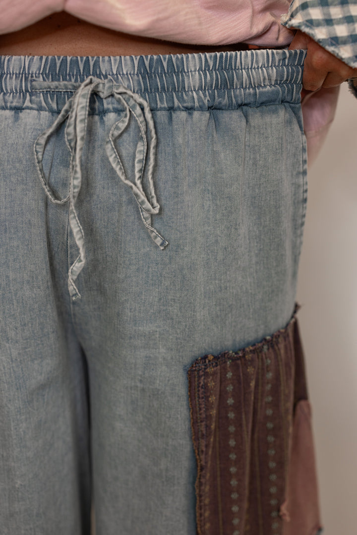 Close-up of a person wearing denim pants with a brown patchwork details and drawstring and elastic waistband