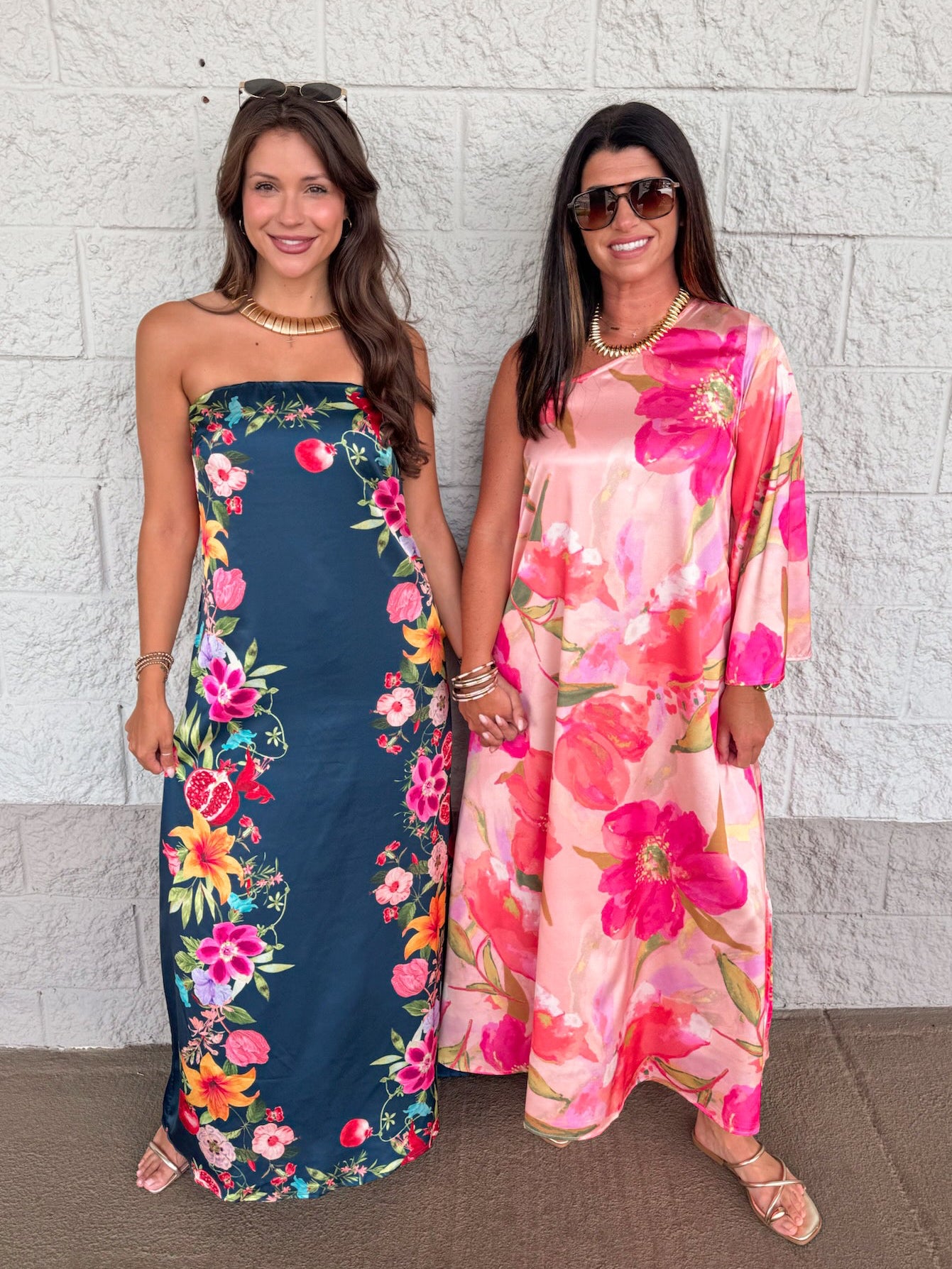 Two women standing side-by-side in floral maxi dresses against a white brick wall. The woman on the left wears a strapless navy blue dress with a colorful floral print, and the woman on the right wears a pink and orange floral kaftan-style dress.